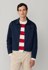 Jeune homme portant une veste zippée bleu marine sur un pull rayé blanc et rouge, avec un pantalon beige, debout devant un fond uni gris clair.