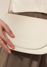 Cremfarbene Leder-Crossbody-Tasche mit einem geschwungenen Deckel und goldfarbener, geprägter Logoplatte, ergänzt durch feine Nähte und ein minimalistisches Design.