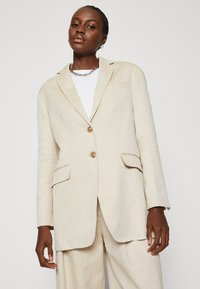 Beige Leinenblazer mit tailliertem Design, einreihiger Knopfschluss, zwei vorderen Taschen und hellbraunen Knopfakzenten.