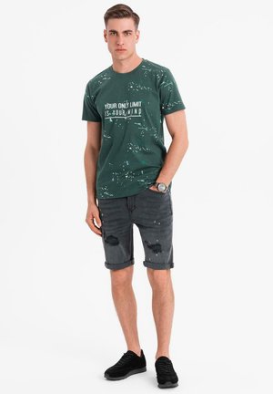 Camiseta de algodón verde con un diseño gráfico en texto; combinada con pantalones cortos de mezclilla grises con detalles desgastados; zapatillas negras.