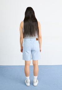Shorts en denim bleu clair avec un détail à la taille, associés à un débardeur gris, des chaussettes blanches et des baskets claires, vus de dos.