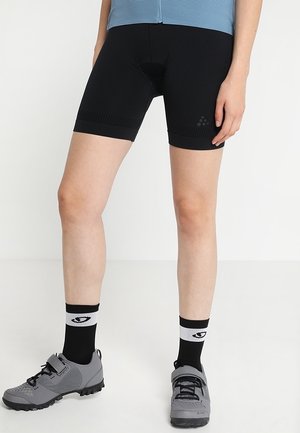 Persoon met zwarte wielershorts, zwart-witte sokken en grijze wielerschoenen, staand op een effen witte achtergrond.