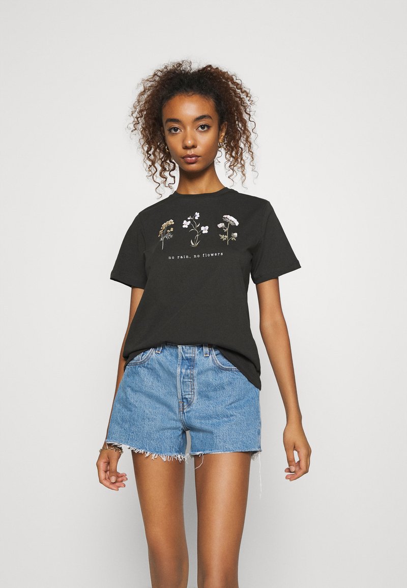 Sort bomulds T-shirt med korte ærmer, der har blomsterbroderi og teksten "ingen regn, ingen blomster" centreret nedenfor. Bæres med blå denim shorts.