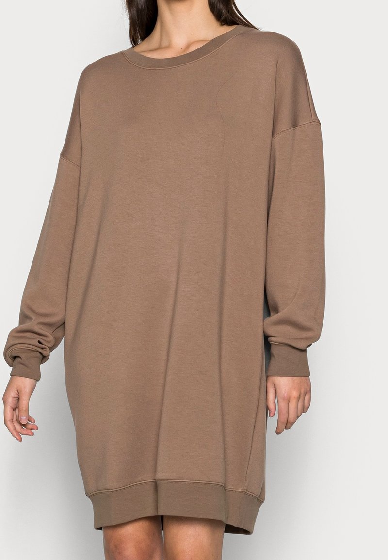 Femme portant une robe sweat ample taupe, à manches longues, longueur genoux, avec un col rond et des poignets côtelés.