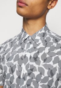 Lyhythihainen paita valkoisena ja mustana, jossa on lehtikuvio ja raidalliset yksityiskohdat. Napillinen kaulus ja sileä kangaspinta.