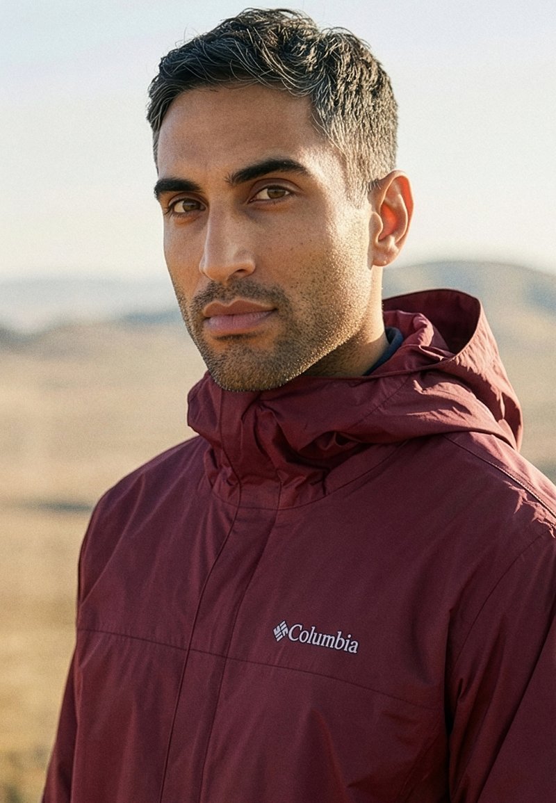 Jonge man met kort donker haar, gekleed in een bordeauxrode Columbia capuchonjas, buiten met een onscherpe natuurlijke landschap op de achtergrond.