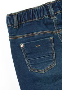 Jean en denim bleu pour enfant avec ceinture élastique et poche arrière présentant un petit rivet en métal en forme d'étoile sur un fond blanc.