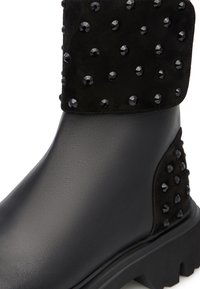 Stivaletto nero con una base in pelle liscia, accentuato da un risvolto in suede nero decorato con borchie nere rotonde, dotato di una suola robusta.