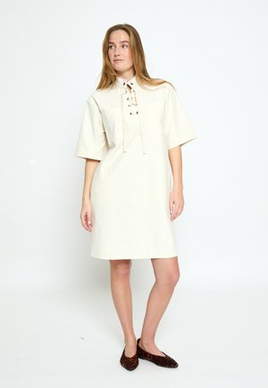 Woman standing wearing a loose cream dress with short sleeves and lace-up neckline, paired with leopard print flats.