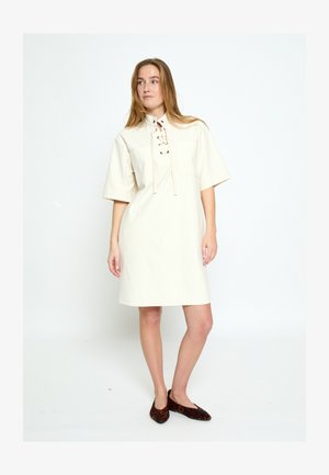 Kobieta stojąca w luźnej kremowej sukience z krótkimi rękawami i wiązanym dekoltem, w połączeniu z balerinami w panterkę.