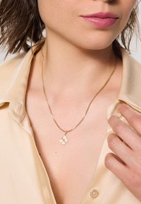 Złoty naszyjnik z łańcuszka z delikatnym charms kwiatowym o teksturowanych płatkach, noszony na gładkiej, kremowej bluzce.