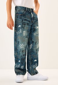 Jeans in denim blu con vestibilità ampia, decorati con vari disegni in stile graffiti in bianco e nero, dotati di cinque tasche e chiusura con bottoni.