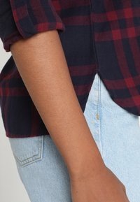 Marineblauwe en bordeaux geruite overhemd met opgerolde mouwen en een gebogen zoom, gemaakt van stof met een soepele textuur. Draagt over lichtblauwe denim jeans.