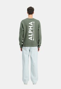Felpa verde oliva con grande testo bianco "ALPHA INDUSTRIES" sulla schiena, polsini a costine e vestibilità rilassata. Indossata con jeans azzurri chiari.