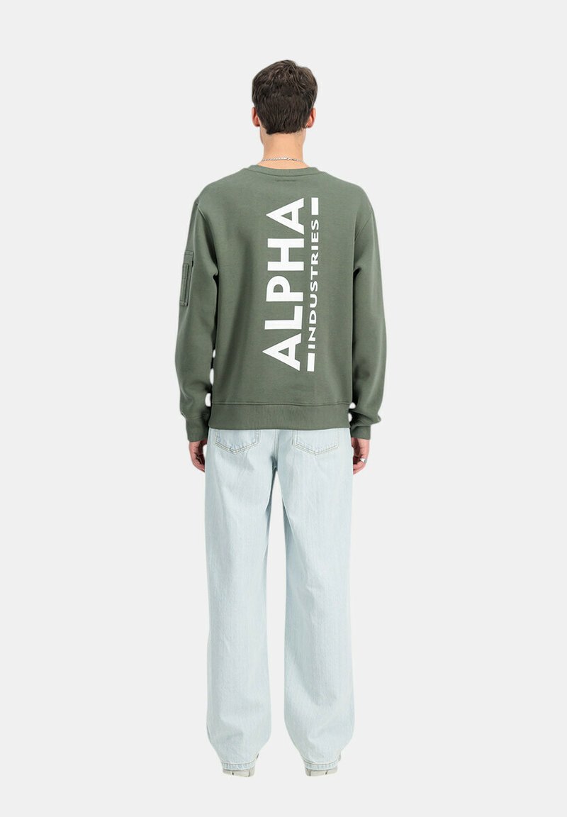 Felpa verde oliva con grande testo bianco "ALPHA INDUSTRIES" sulla schiena, polsini a costine e vestibilità rilassata. Indossata con jeans azzurri chiari.