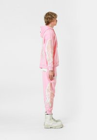 Sudadera con capucha rosa y pantalones de chándal que presentan diseños de alas bordados, confeccionados en un tejido suave. El modelo lleva botas blancas de suela gruesa.