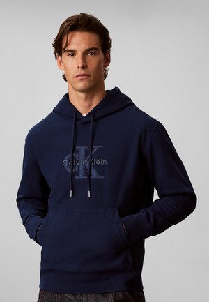 Uomo che indossa una felpa con cappuccio blu navy Calvin Klein con tasca frontale e coulisse, in piedi davanti a uno sfondo chiaro e semplice.