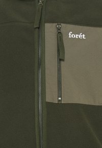 Primo piano di una giacca di pile verde oliva con zip verticale e tasca sul petto con chiusura zip, caratterizzata da un ricamo bianco con la scritta "forét".