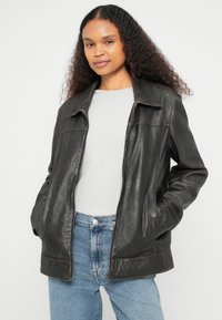 Mustast nahast jakk, millel on tõmblukuga sulgemine, kraega disain ja peen läige. Kandmiseks heledast hallist kampsunist ja sinisest teksastest üle.