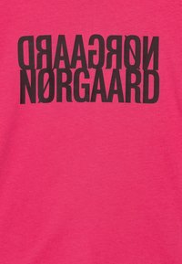 Bavlněné tričko v jasně růžové barvě s velkým, tučným černým textem: "NØRGAARD" a "NØRGAARD" vytištěným vzhůru nohama. Měkká textura, volný střih.