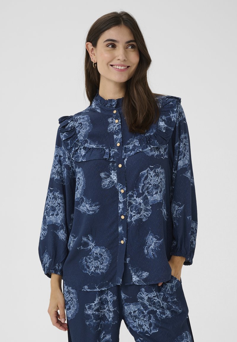 Mörkblå blus med blommönster, volangkrage, långa ärmar och knäppning fram. Lätt tyger med en slät yta.