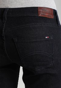 Jeans de denim oscuro con corte ajustado, que presentan una etiqueta de cuero marrón con "Tommy Jeans" y dos bolsillos traseros con costuras a tono.