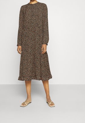 Frau trägt ein langärmliges, schwarzes Midikleid mit Blumenmuster und beige flache Sandalen, steht vor einem schlichten, hellen Hintergrund.