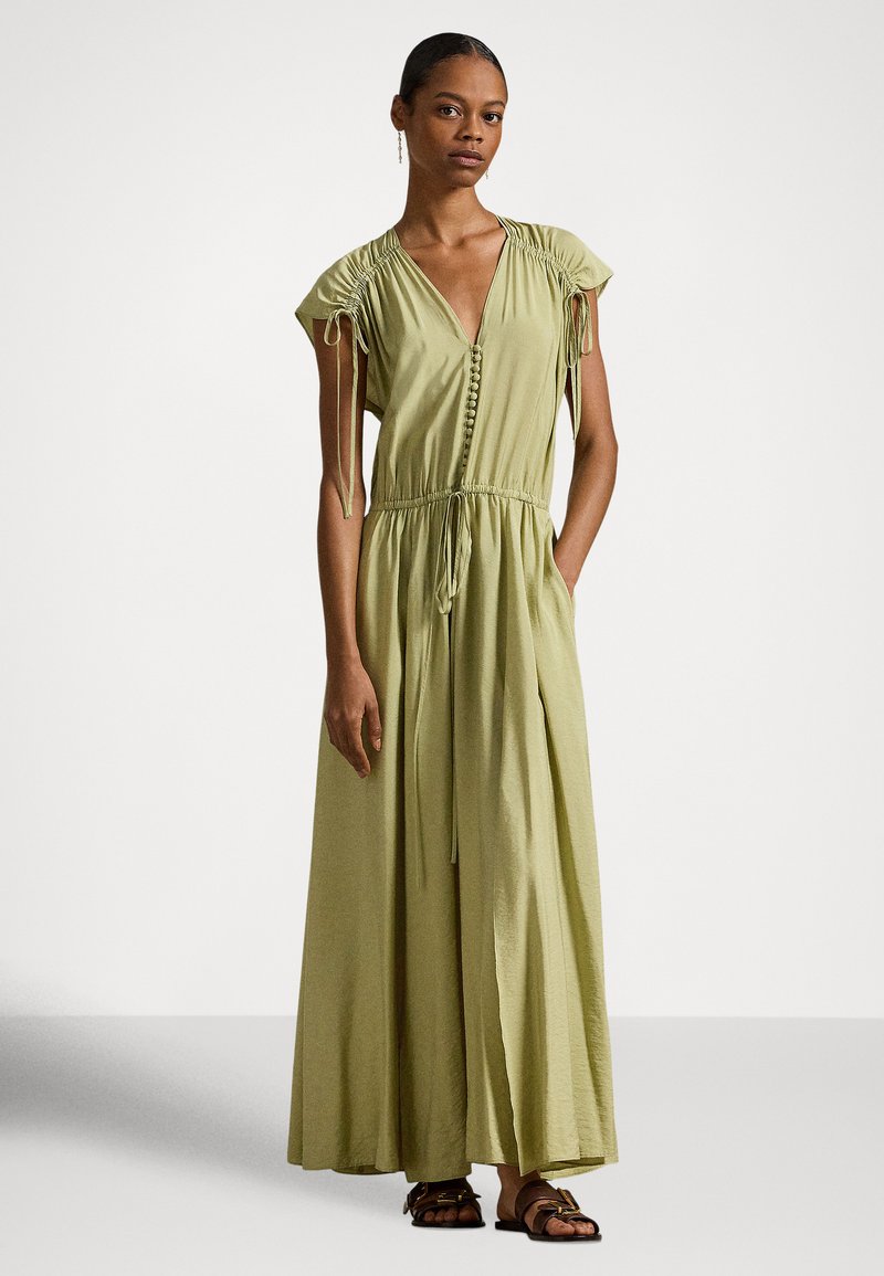 Γυναίκα που φοράει μακρύ ελιά πράσινο φόρεμα με κοντά ρυτιδωμένα μανίκια, εμπρόσθιο μέρος με κουμπιά και κορδόνι στη μέση, στέκεται με το ένα χέρι στην τσέπη.