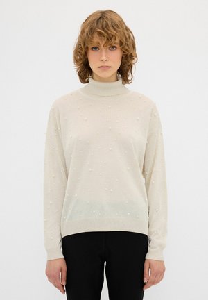 Cremefarvet turtleneck-sweater med en løs pasform, tekstureret strik, dekoreret med små perle-lignende accenter i stoffet. Sorte bukser er vist.