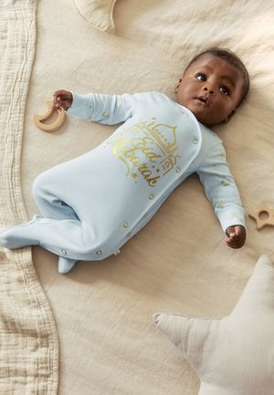 Baby, der ligger på beige tæppe iført lyseblå heldragt med fødder og gyldent "Eid Mubarak"-tekst, holder et trælegetøj formet som en halvmåne, nær en stjernepude.