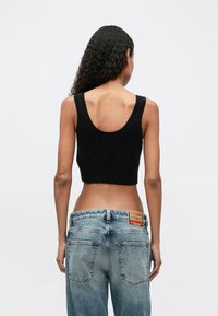 Žena s dugom kovrčavom kosom, odjevena u crni bezrukavni crop top i široke plave traperice, prikazana sa stražnje strane na jednoličnoj pozadini.
