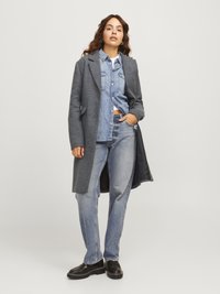 Cappotto di lana grigio con tasche laterali indossato sopra una camicia di jeans azzurri chiari e jeans. Scarpe nere con suola spessa. Sfondo semplice.