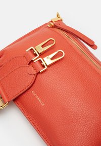 Bolso de cuero naranja con superficie texturizada, herrajes dorados, dos cierres a presión y un compartimento con cremallera. Nombre de la marca visible.