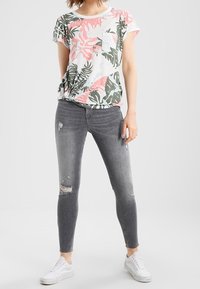 Vit t-shirt med rosa och grön tropisk bladmönster, fickdetalj; kombinerad med mörkgrå rivna skinny jeans och vita sneakers.