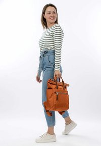 Zaino arancione in nylon con cima arrotolata, tasca con zip e spallacci doppi, abbinato a una maglietta a maniche lunghe a righe e jeans a vita alta.