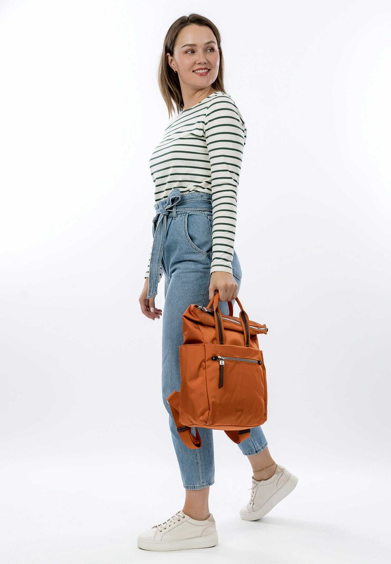 Zaino arancione in nylon con cima arrotolata, tasca con zip e spallacci doppi, abbinato a una maglietta a maniche lunghe a righe e jeans a vita alta.