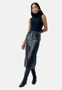 Top negro sin mangas combinado con una falda midi negra de cuero sintético de talle alto que presenta una abertura frontal y un acabado brillante, complementado con botas hasta la rodilla.
