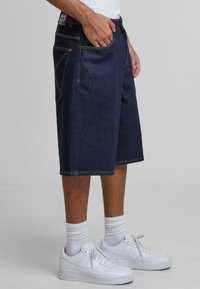 Pantalones cortos de mezclilla en azul oscuro, con bolsillos laterales y traseros, costuras amarillas y un ajuste holgado. Llevados con zapatillas blancas y calcetines de caña.