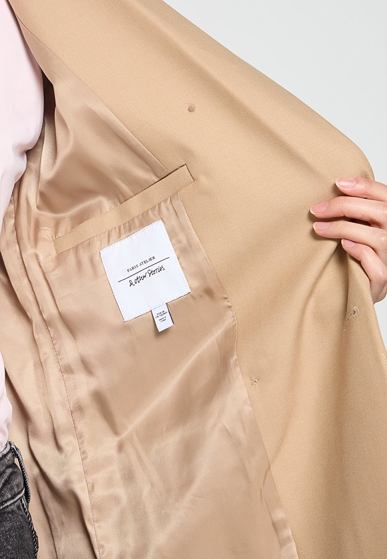Beiger maßgeschneiderter Blazer mit Satin-Textur, ausgestattet mit einer Innentasche und Etikett. Das Futter ist in einem hellen Cremeton.
