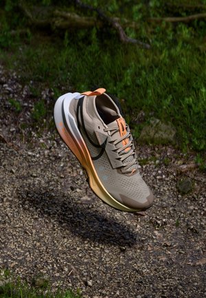 Chaussure de course beige et orange avec un logo noir en forme de virgule, en l'air au-dessus d'un chemin de gravier et d'herbe verte dans un environnement extérieur.
