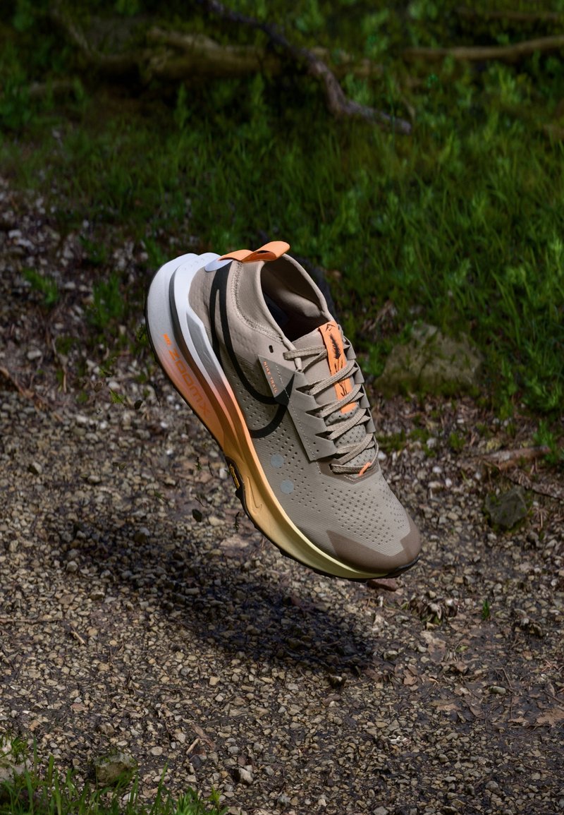 Chaussure de course beige et orange avec un logo noir en forme de virgule, en l'air au-dessus d'un chemin de gravier et d'herbe verte dans un environnement extérieur.