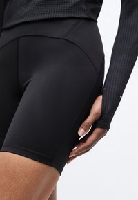 Κοντινή λήψη ενός ατόμου που φοράει μαύρο αθλητικό σορτς και μακρυμάνικη μπλούζα με υφή, με το χέρι να ακουμπά κοντά στο μπράτσο.