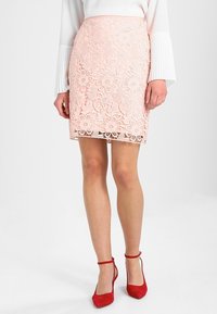 Pink lace skirt with floral patterns, lined interior, and scalloped hem, paired with red strap heels. Smooth texture and fitted design.