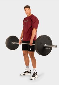 Bordeaux oversized t-shirt, zwarte shorts, houdt een halterstang vast met zwarte gewichten. Zwarte sport schoenen en witte sokken met strepen.