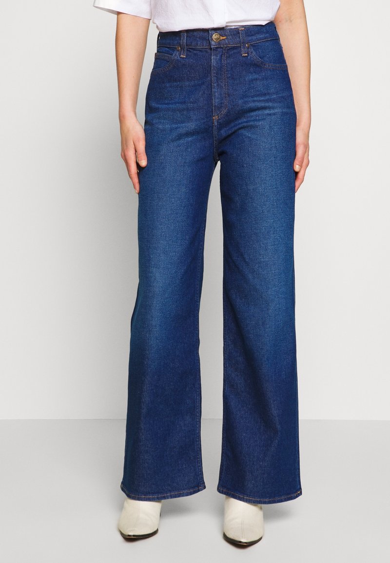 Personne portant un jean bleu foncé taille haute à jambes larges, associé à des chaussures blanches et un haut blanc, debout devant un fond uni.