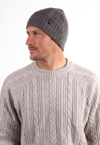 Bonnet en tricot gris avec un motif côtelé, porté par une personne dans un pull léger en tricot torsadé. Le pull présente des motifs verticaux texturés.