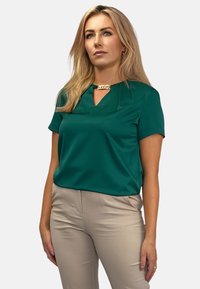 Niebieska bluzka z krótkim rękawem i dekoltem w kształcie litery V z złotym akcentem. Gładki materiał; w połączeniu z beżowymi dopasowanymi spodniami. Prosty design bez wzorów.