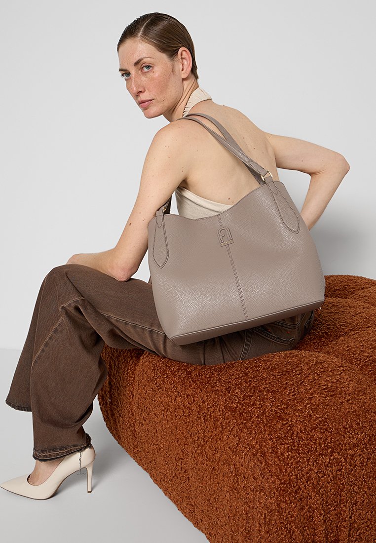 Jasnoszara skórzana torba hobo o teksturowanej powierzchni, z dwiema zaokrąglonymi uchwytami i subtelnym akcentem logo. Umiejscowiona obok miękkiej, teksturowanej powierzchni.