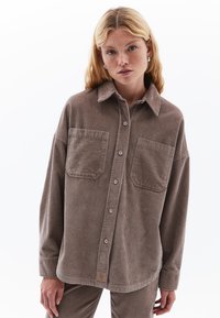 Bruine ribfluwelen blouse met een knoopsluiting aan de voorkant, kraag, twee voorzakken en een label aan de zoom. Wijde mouwen en ribbelstructuur.