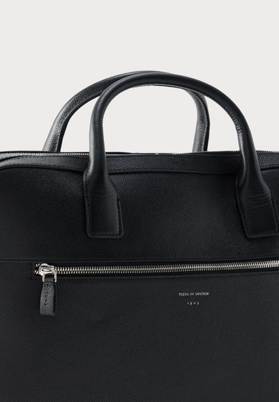 Μαύρη δερμάτινη τσάντα με υφή, διπλές λαβές, μπροστινή τσέπη με φερμουάρ και ανάγλυφο λογότυπο "Tiger of Sweden 1903" κάτω από το φερμουάρ.